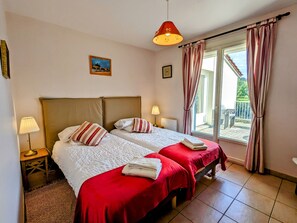 A double room with two beds, each with a red bedspread and white pillows, a small bedside table with a lamp, a painting on the wall, and a view of the outside through a sliding glass door. - Riverside Retreat: Heated Pool, Tennis, 5 Bedrooms, Private Parking (Haironville)
