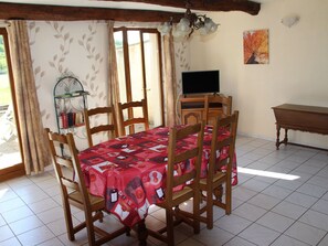 A dining area with a table covered in a red tablecloth, wooden chairs, a television on a wooden stand, and a painting on the wall.