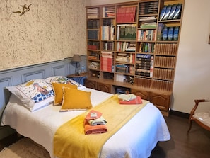 A bedroom with a bed, pillows, a bedside table, a lamp, a bookshelf, and a chair.