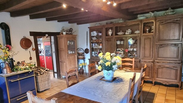 Une salle à manger traditionnelle avec une table en bois, des chaises et un vase de fleurs. On y trouve des meubles contenant de la vaisselle et, au fond, un appareil électroménager rouge.