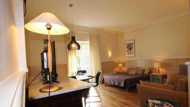 A hotel room with a bed, a desk with a computer, a chair, a small table with magazines, and a lamp.