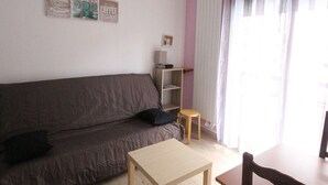 A living room with a brown sofa, a small wooden table, a chair, a shelf with items, and a window with curtains.