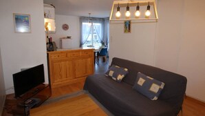 A cozy living room with a sofa, a coffee table, and a TV. There's a kitchen area with a fridge and a dining table. - Central Apartment near Spa, 2 Bedrooms, Sleeps 6, Parking Included (Bagnères-de-Luchon)