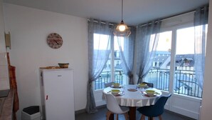 A dining area with a table set for four, a refrigerator, a clock, and a view of the outside through large windows. - Central Apartment near Spa, 2 Bedrooms, Sleeps 6, Parking Included (Bagnères-de-Luchon)