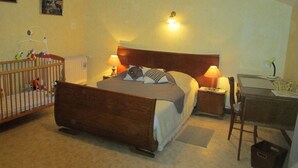 A bedroom with a wooden bed, two bedside lamps, a crib with toys, a desk with a chair, and a radiator. - Charming Rural Retreat with Modern Amenities near Verdun – Pets Welcome! (Bréhéville)