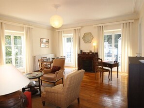 A spacious living room with a fireplace, wooden flooring, and large windows offering a view of the outdoors.