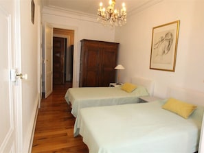 Une chambre avec deux lits, une armoire en bois, un lustre et une œuvre d’art encadrée accrochée au mur.