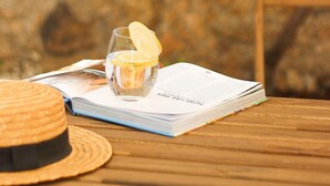 Una mesa de madera con un libro, un vaso de agua con una rodaja de limón y un sombrero de paja.