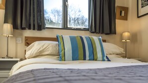 A neatly made bed with a striped pillow, a bedside lamp, and a window with curtains. - Rock Cottage - 3 Bedroom Holiday Home (Fishguard)