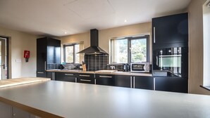 A modern kitchen with black cabinets, a large island, a built-in oven, and a window overlooking greenery. - Rock Cottage - 3 Bedroom Holiday Home (Fishguard)