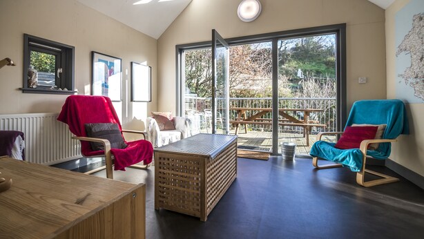 A modern living room with a wooden coffee table, two rocking chairs, a red and a blue chair, and a large window offering a view of the outdoors. - Rock Cottage - 3 Bedroom Holiday Home (Fishguard)