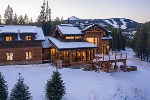 A large wooden lodge with multiple levels, a covered deck, and a view of snow-covered mountains. - Wraparound Porch | Hot Tub | Sauna (Big Sky)