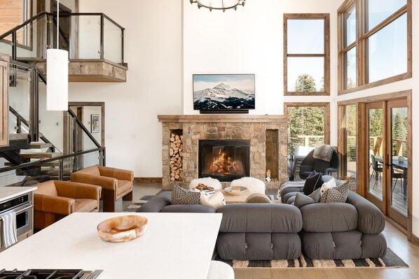 A spacious living room with a fireplace, a sofa set, a dining area with a white table, and a staircase with glass railings. - Wraparound Porch | Hot Tub | Sauna (Big Sky)