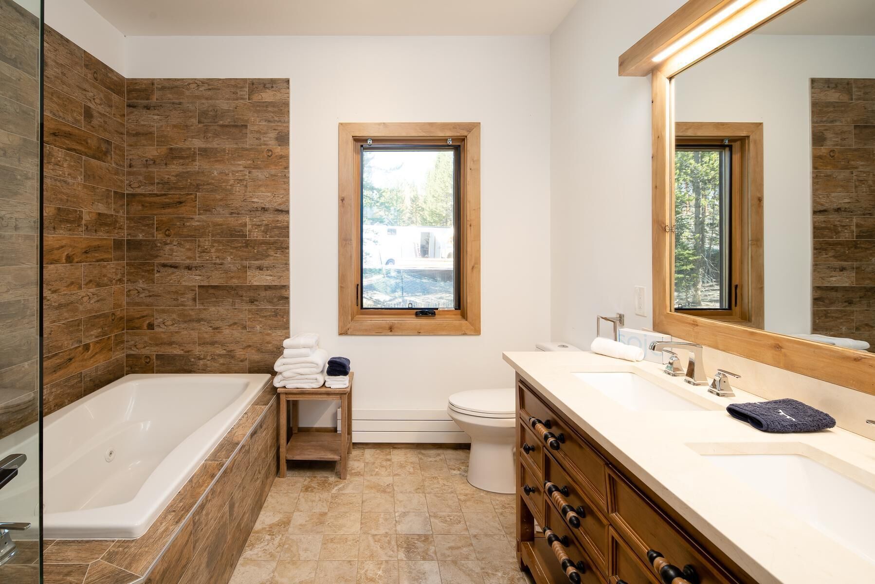 A modern bathroom with a wooden wall, a large window, a bathtub, a toilet, and a vanity with a mirror.