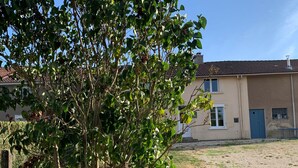 A building with a flat roof and a blue door, surrounded by a grassy area and a tree with green leaves. - Charming Renovated Home near Verdun with Modern Amenities, Terrace, and Garden (Forges-sur-Meuse)