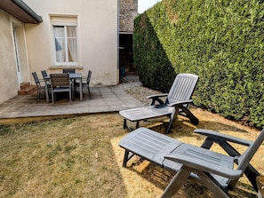 An outdoor patio with a table and chairs, a lounge chair, and a well-maintained hedge.