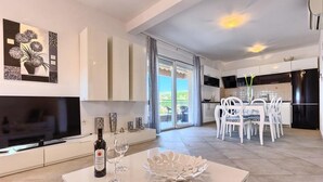A modern kitchen with white cabinets, a dining area with white chairs, and a large TV on the left.