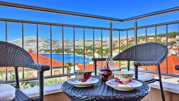 A balcony with wicker chairs and a table, overlooking a coastal town with red-roofed buildings and a blue sea.