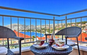 A balcony with wicker chairs and a table, overlooking a coastal town with red-roofed buildings and a blue sea. - Three Bedroom Apartment with Pool (Okrug Gornji)
