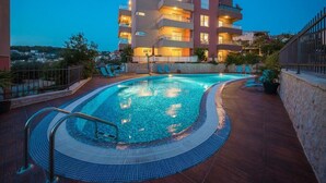 A swimming pool with a tiled deck, surrounded by a stone wall and a building with multiple balconies.