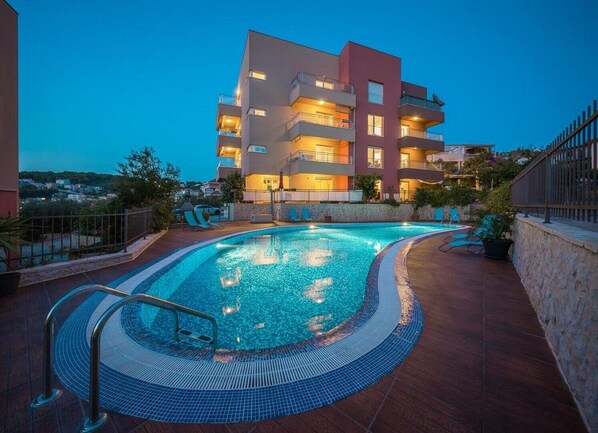A swimming pool with a tiled deck, surrounded by a stone wall and a building with multiple balconies.