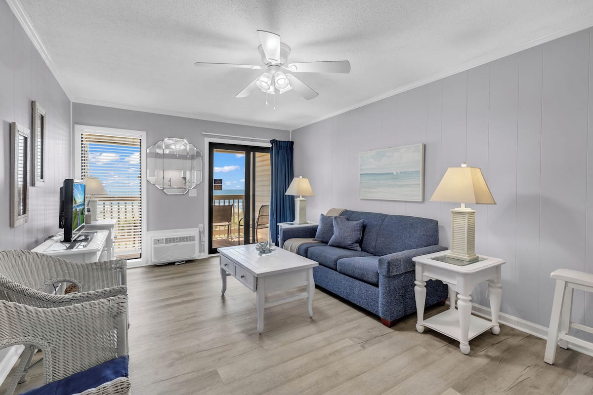 A living room with a white ceiling fan, a blue sofa, a white coffee table, and a white side table with a lamp.