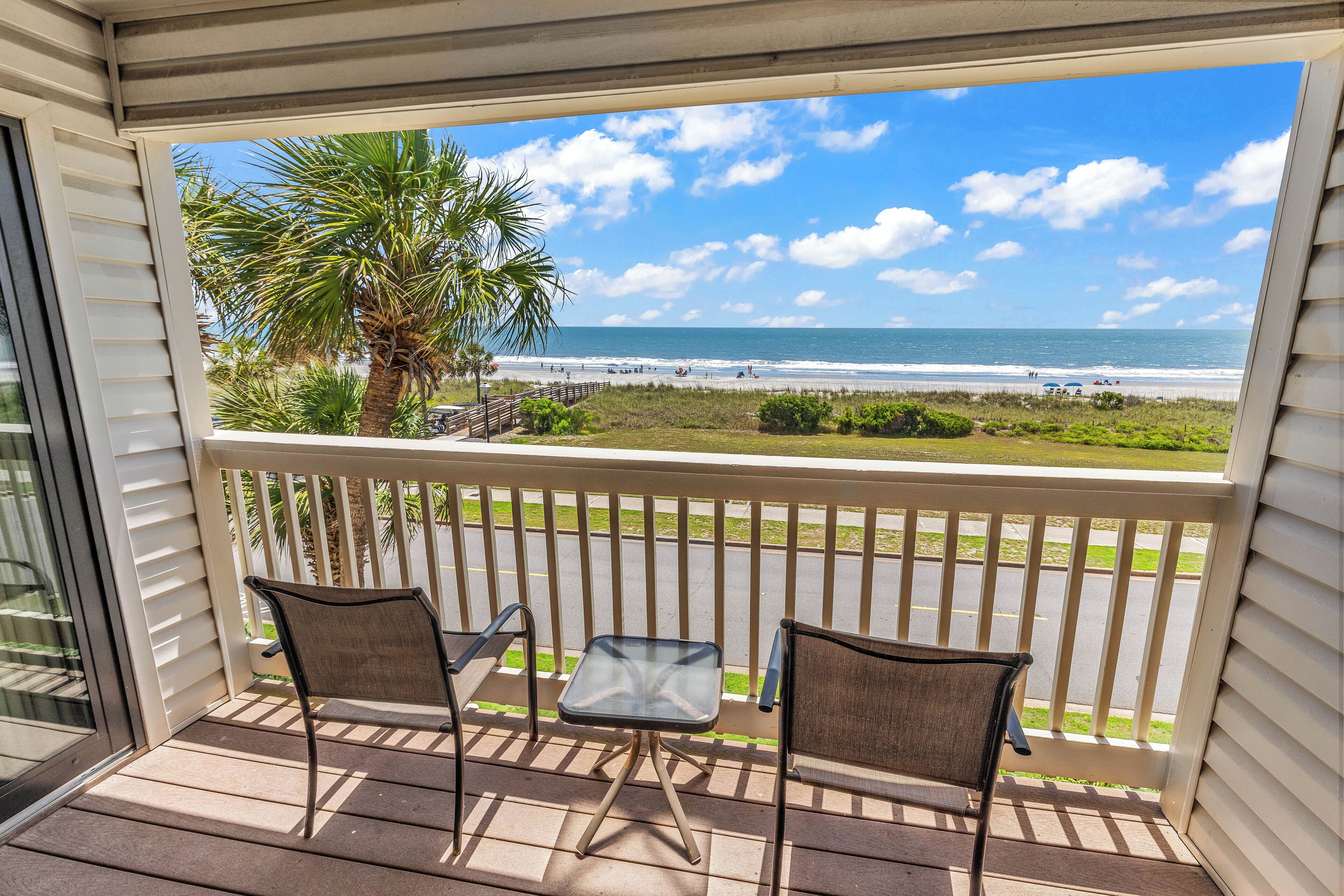 A balcony with two chairs and a small table, overlooking a beach with palm trees and a clear sky.