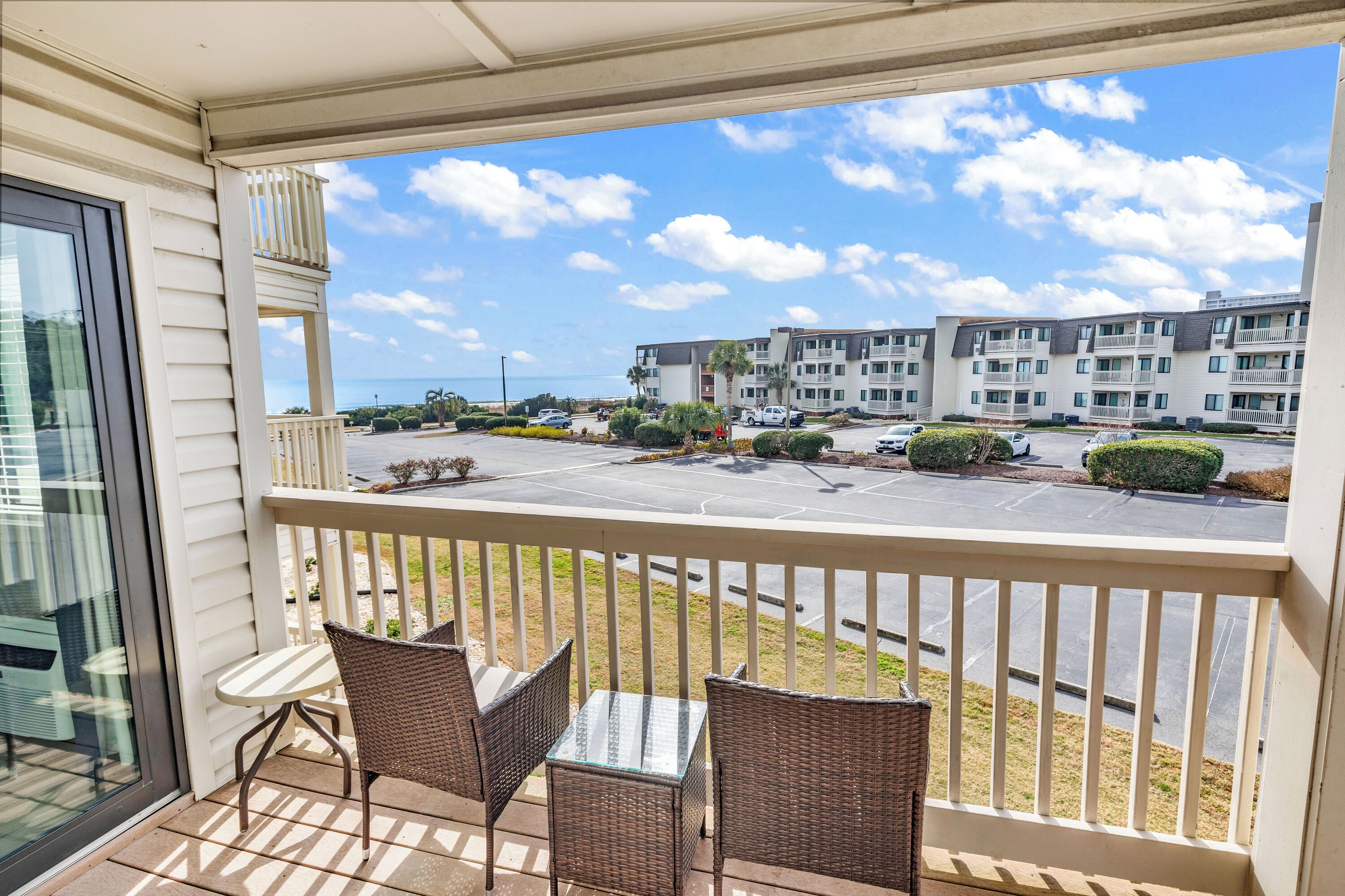A balcony with wicker chairs and a table, overlooking a parking lot and a building with multiple balconies and windows.