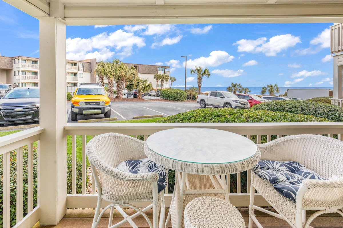A balcony with wicker chairs and a glass table, overlooking a parking lot and buildings with a view of the ocean.