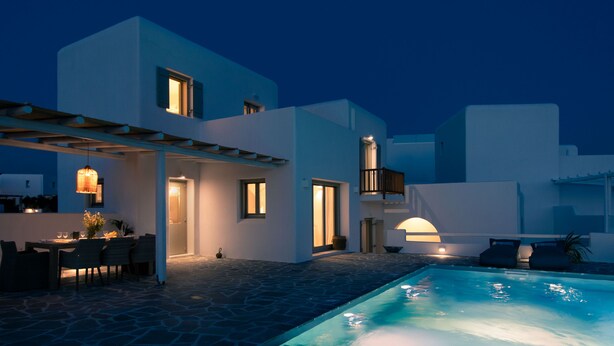 A modern outdoor pool area with stone flooring, a dining setup, and a well-lit building with multiple windows. - Villa ILI - An Uncompromised luxurious escape  (Naxos)