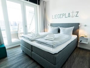 Un lit bien fait, avec du linge de lit blanc et des oreillers, une tête de lit rembourrée de couleur grise et une étagère fixée au mur, sur laquelle se trouvent un livre et un téléphone.