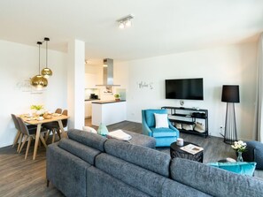 Un salon moderne avec un canapé d’angle gris, un fauteuil bleu et un lampadaire noir. On y trouve également un coin repas avec une table et des chaises en bois, et une cuisine à l’arrière-plan.
