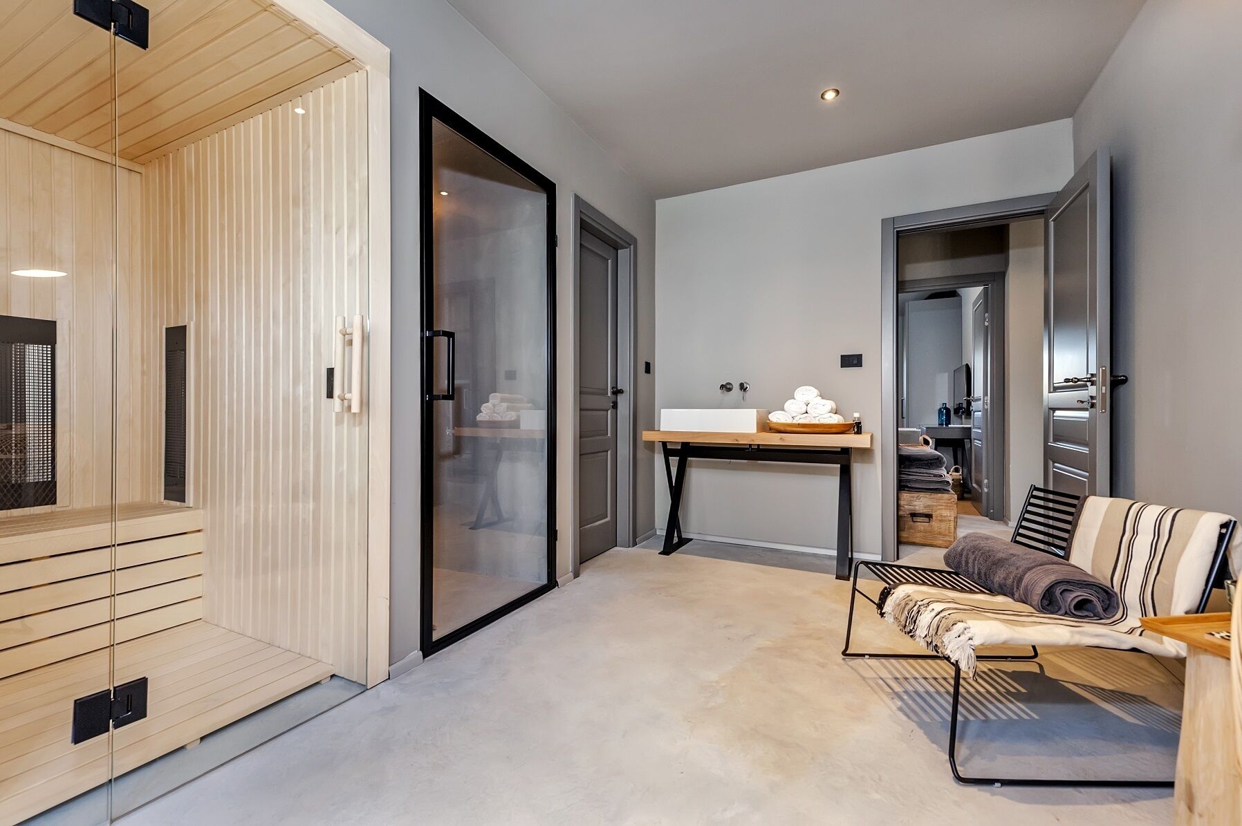 A modern hotel room with a sliding glass door leading to a bathroom, a wooden sauna, a small table with towels, and a sofa with a striped cushion.