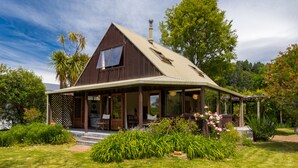 Une maison en bois à deux étages, dotée d’un grand porche à l’avant, d’une cheminée et d’un jardin où poussent diverses plantes et fleurs.