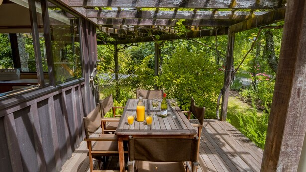 Une terrasse en bois avec une table dressée pour le repas, entourée de chaises, et une pergola au-dessus.