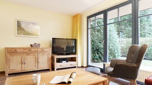 A room with a wooden table, a television, a sofa, and a window with a view of greenery. - Papillon Whg. 06/1, Germany (Ostseebad Boltenhagen)