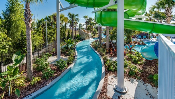 Ein Wasserpark mit einem gewundenen Pool, grünen und blauen Rutschen, Palmen und blauem Himmel.