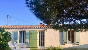 Una casa de un solo piso con techo de tejas, contraventanas verdes y un pequeño porche con dos sillas blancas.