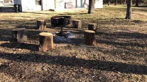 Un espacio al aire libre rústico con un brasero rodeado de troncos de madera, un pequeño edificio con techo inclinado y una bandera.