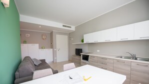 Een moderne keuken met witte kasten, een magnetron en een spoelbak. Er is een zithoek met een witte tafel en een grijze bank.