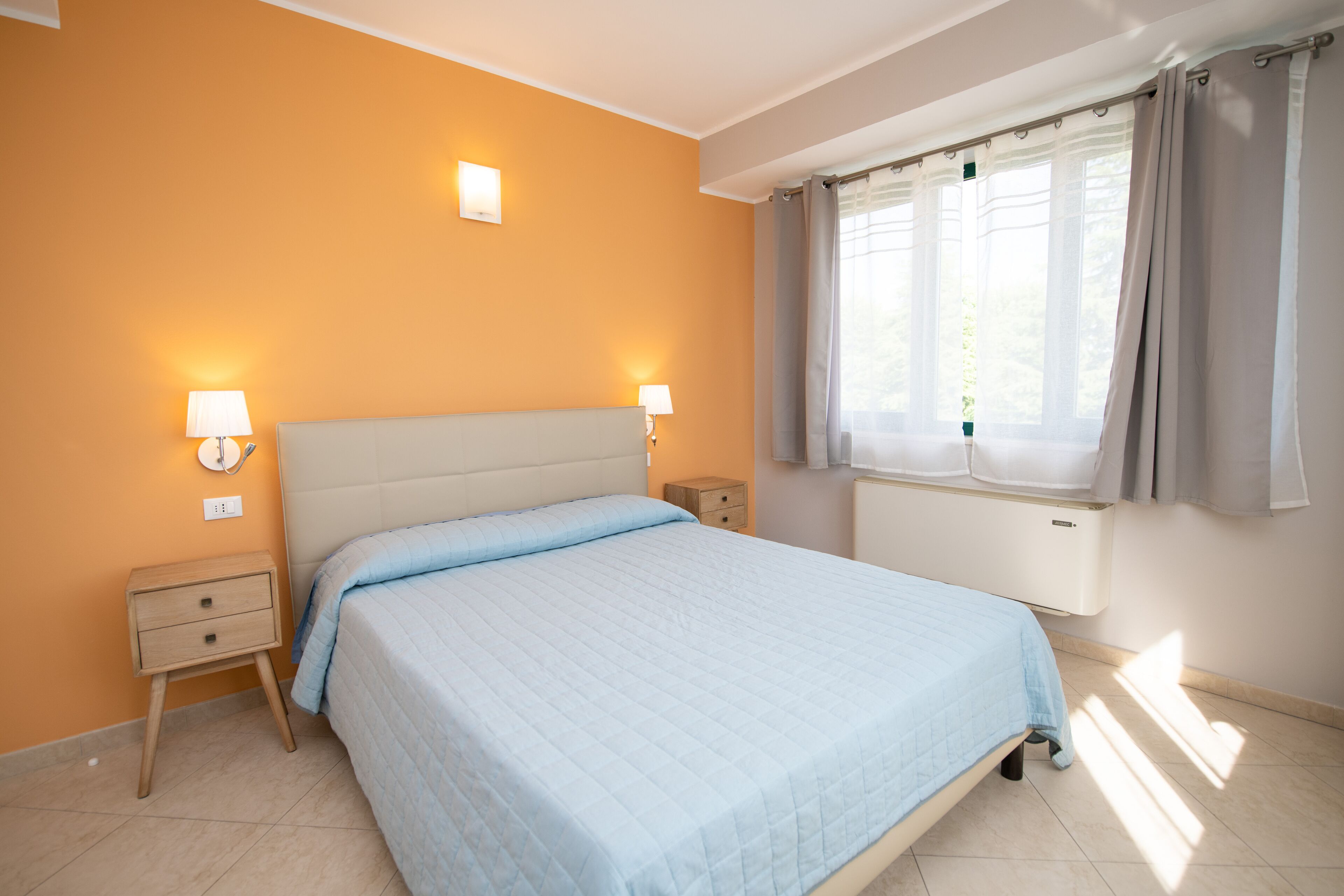 Un dormitorio con cama, mesitas de noche, lámpara de pared y cortinas en la ventana.