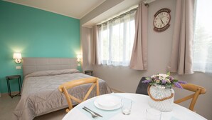 Een slaapkamer met een bed, nachtkastjes, een ronde eettafel met stoelen, een vaas met bloemen en een wandklok.