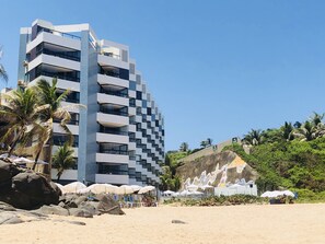 A modern hotel with a unique geometric facade, located adjacent to a sandy beach with palm trees and a rocky cliff.