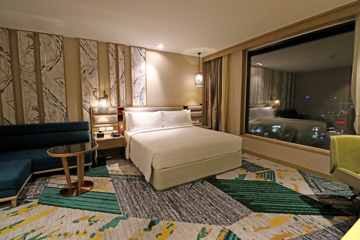 A hotel room with a bed, a sofa, a small table, a nightstand, a lamp, and a large window with a city view.