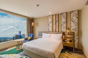 Habitación de hotel con una cama grande, una silla, una mesita y vistas al horizonte de la ciudad.