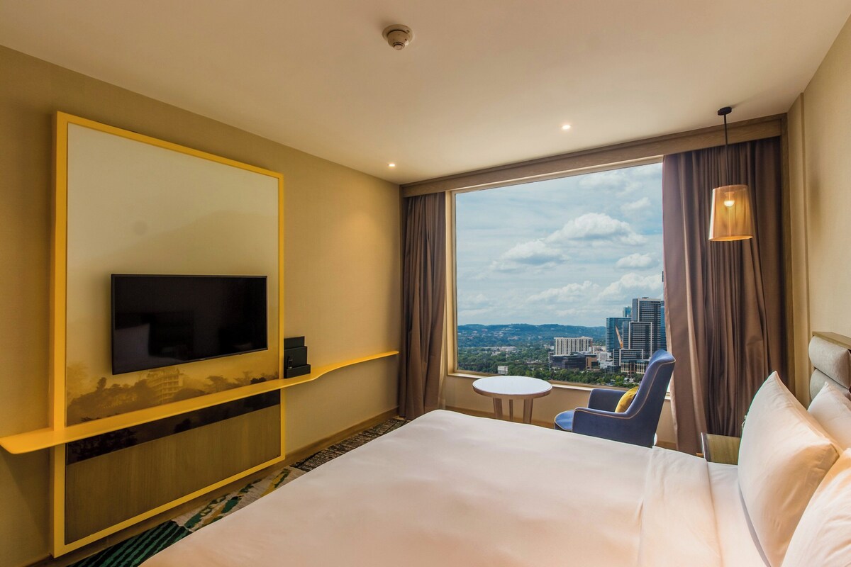 A hotel room with a large bed, a television mounted on a wall, a small round table, a chair, and a view of a cityscape through a large window.