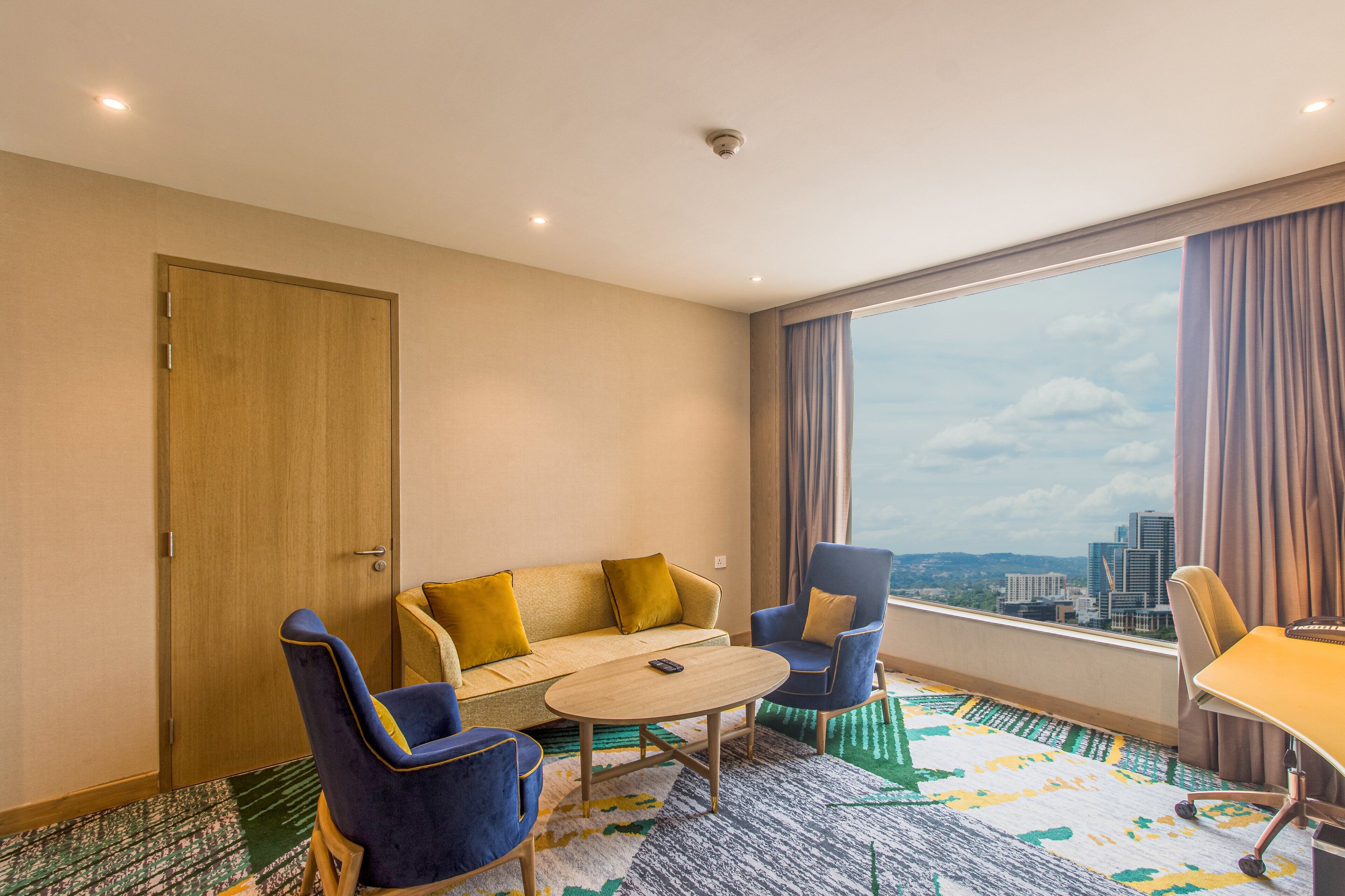 A hotel room with a sofa, two armchairs, a small table, and a large window with a city view.