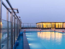 Una piscina en la azotea con barandillas de vidrio, un área de descanso con luces colgantes y vistas a la ciudad.