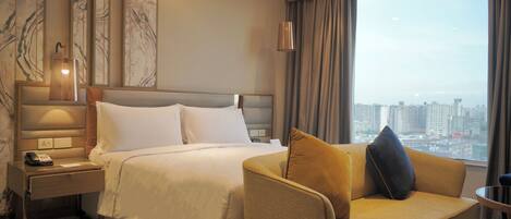 Un cuarto de hotel con una cama, un escritorio con teléfono, una silla, una lámpara y vistas a un paisaje urbano a través de un ventanal grande.