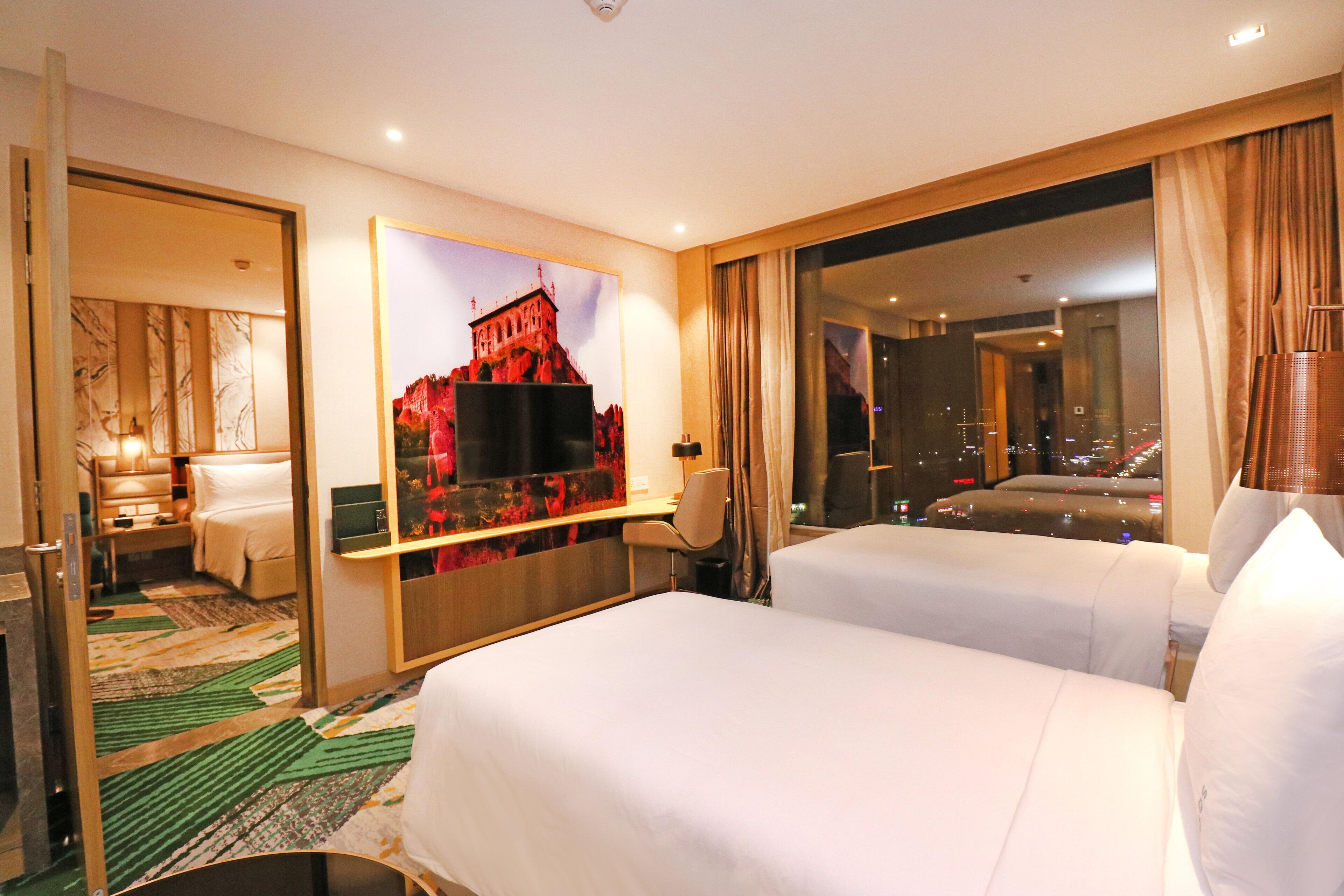 A hotel room with two beds, a large window with a city view, a flat-screen TV, and a decorative wall piece.
